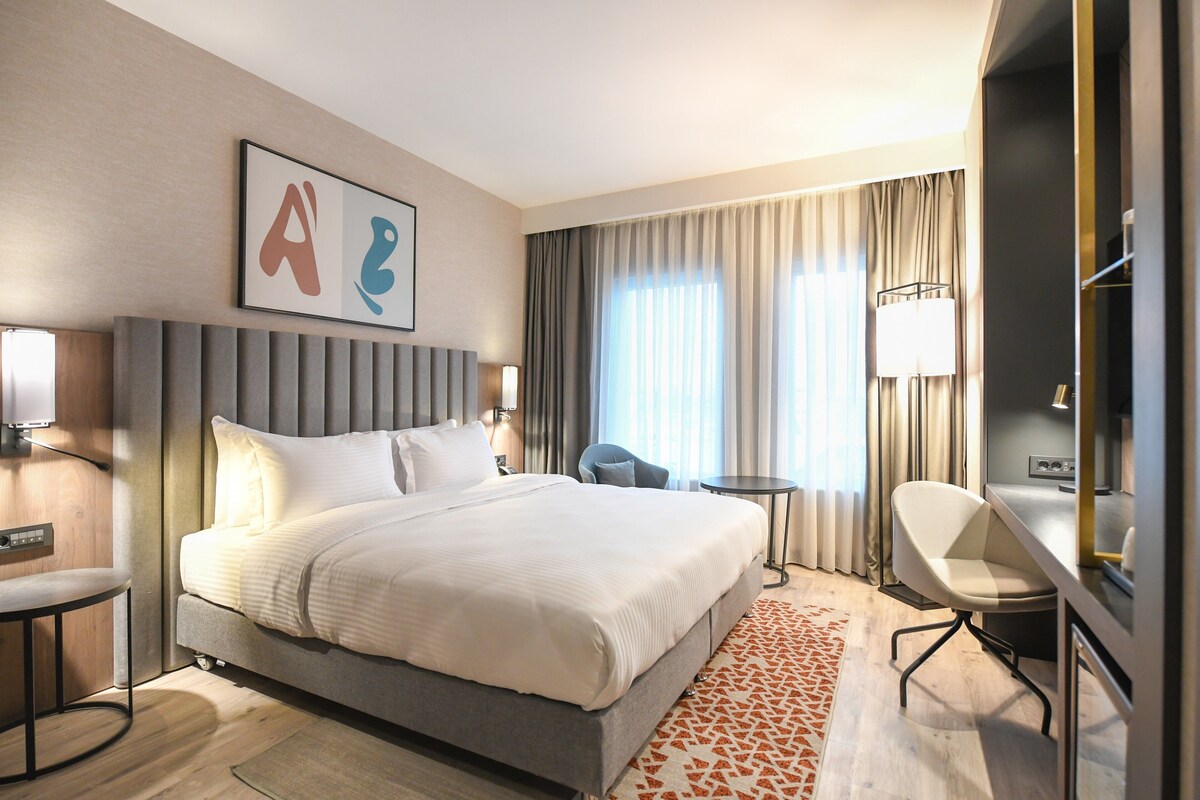 A modern hotel room with a large bed, a desk, and a chair. There is a window with curtains, a wall-mounted artwork, and a lamp.