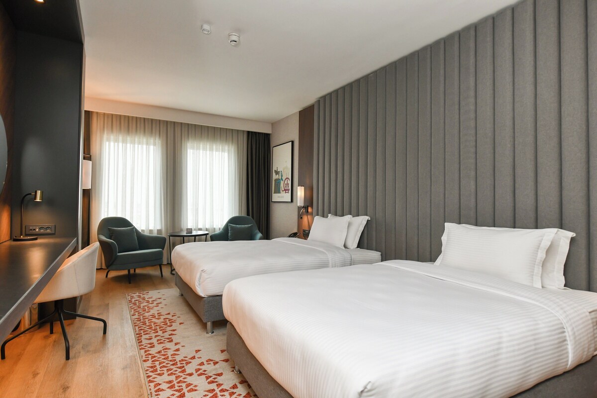 A hotel room with two beds, a desk, a chair, and a large window with curtains.