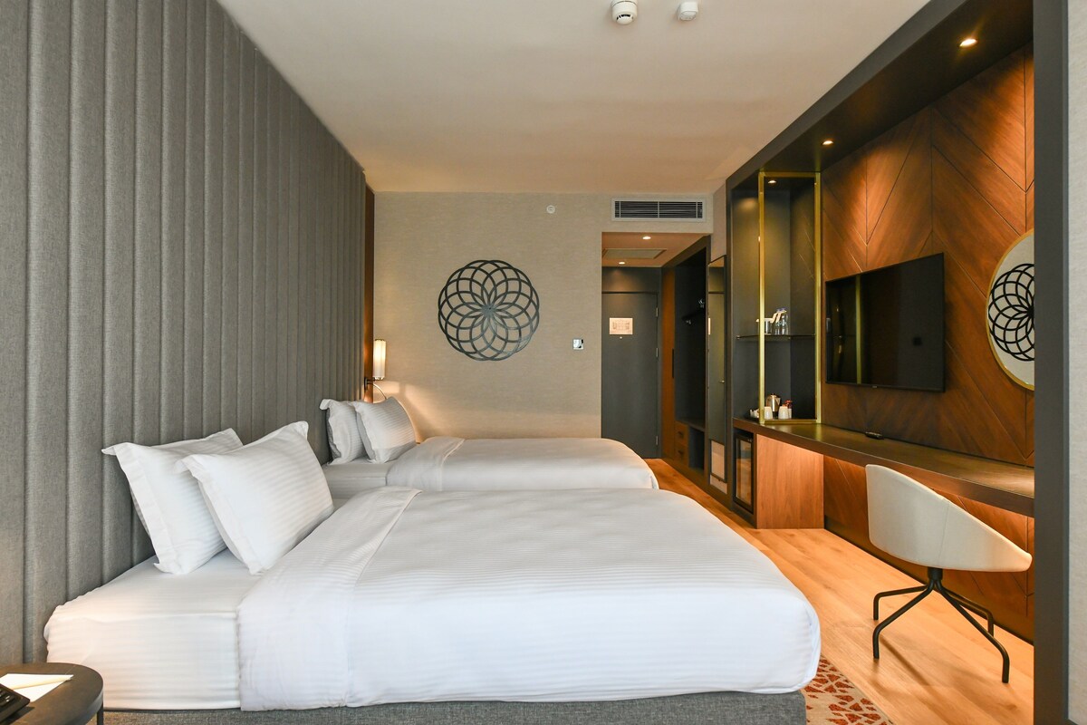 A modern hotel room with two beds, a desk, and a TV.