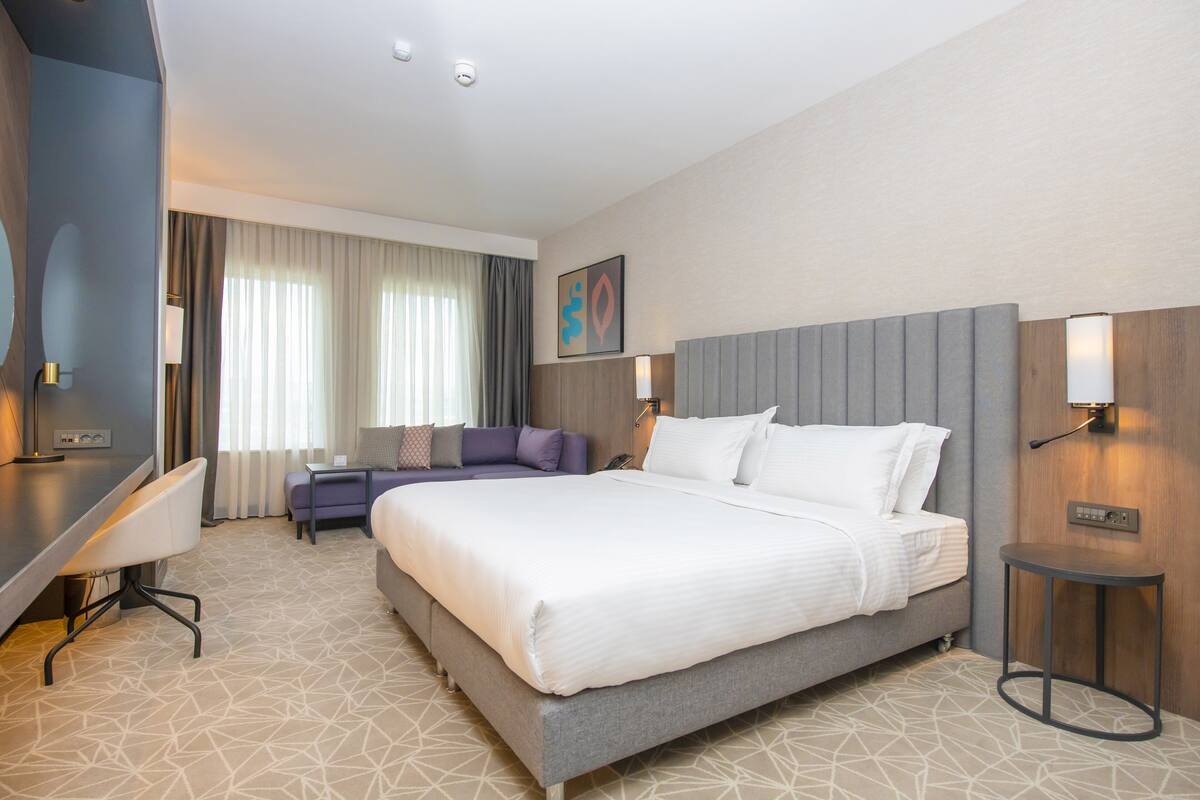 A hotel room with a large bed, a desk, a chair, a sofa, and a small table.