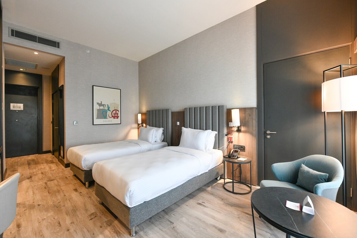 A hotel room with two beds, a sitting area with a table and chair, and a door with a light switch.