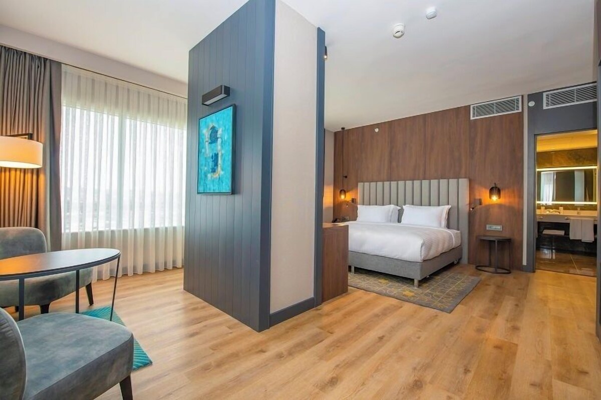 A modern hotel room with a bed, bedside table, lamp, and a view of the city.