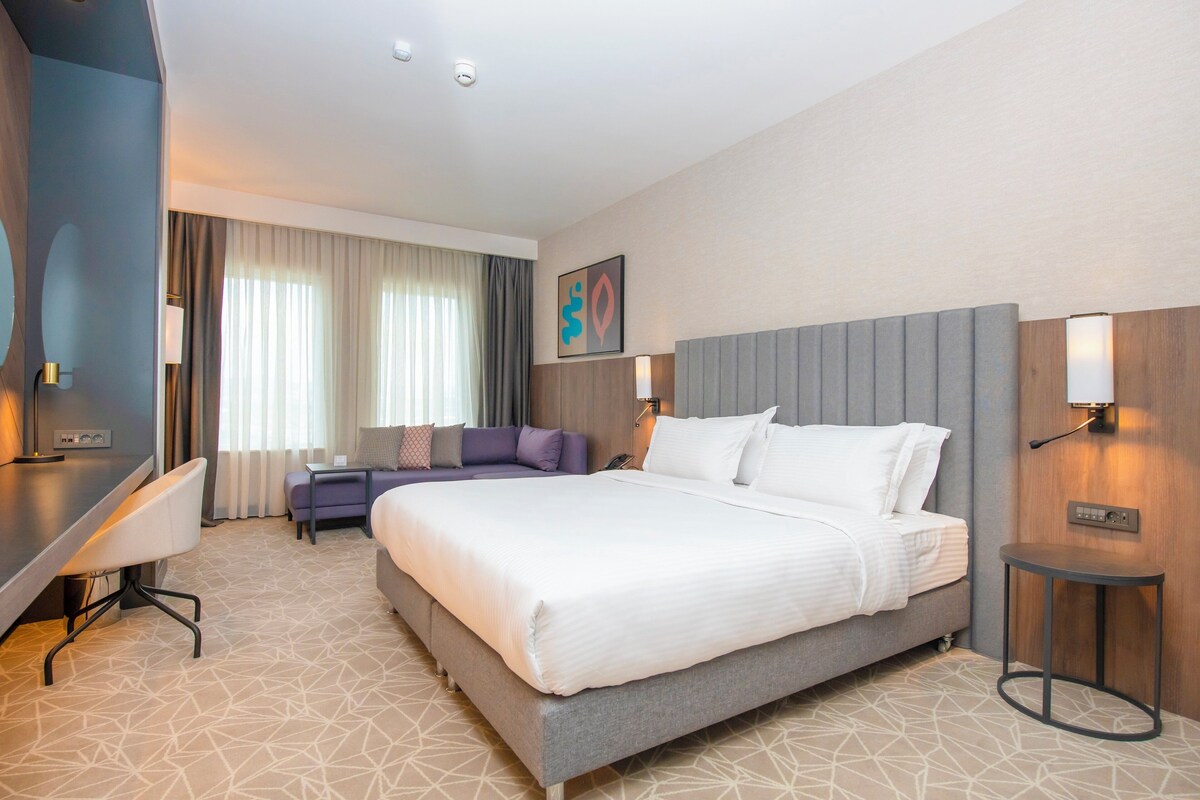 A hotel room with a large bed, a desk, a chair, a sofa, and a small table.