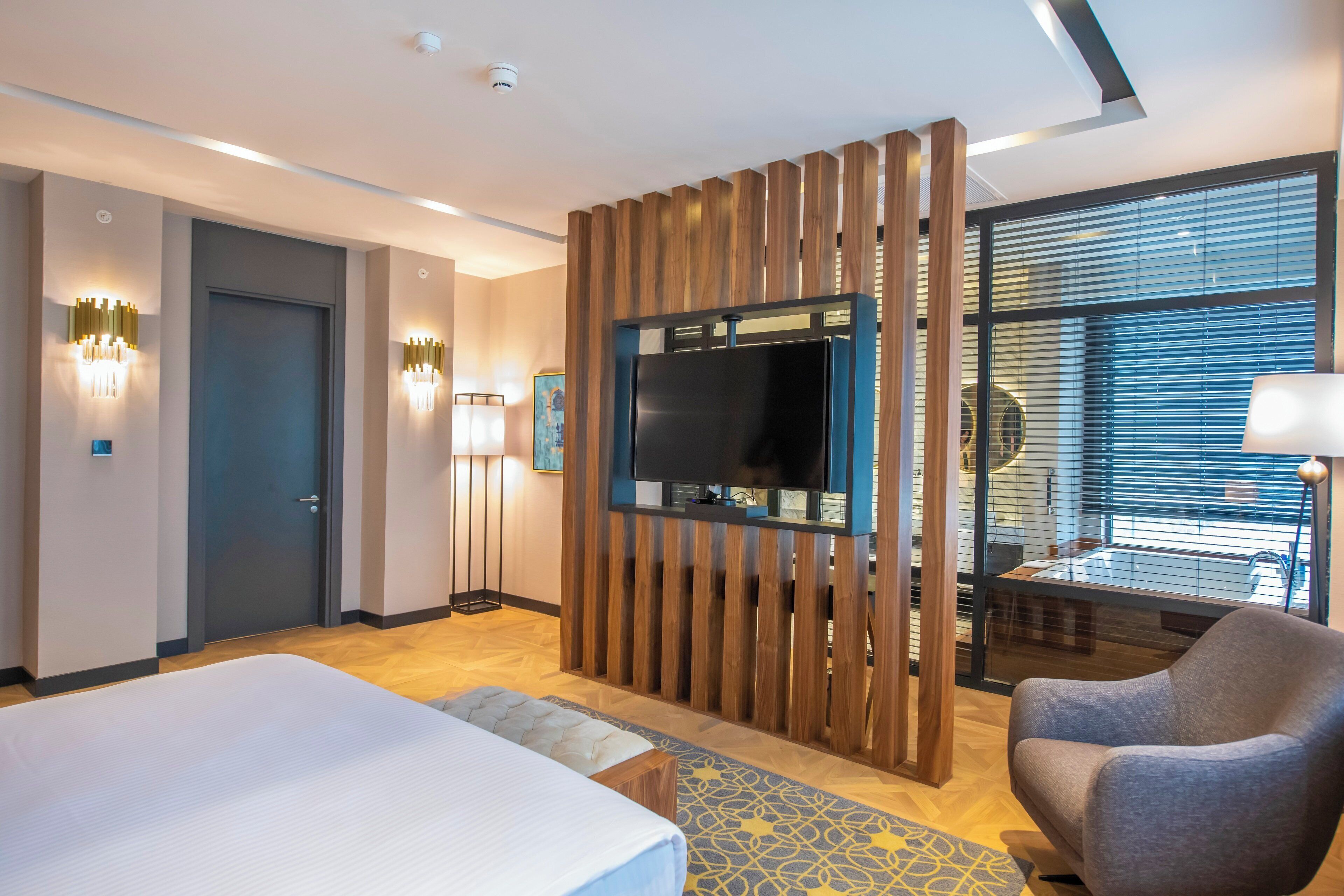 Habitación de hotel con cama, televisor montado en un panel de madera, una silla y ventana con persianas.