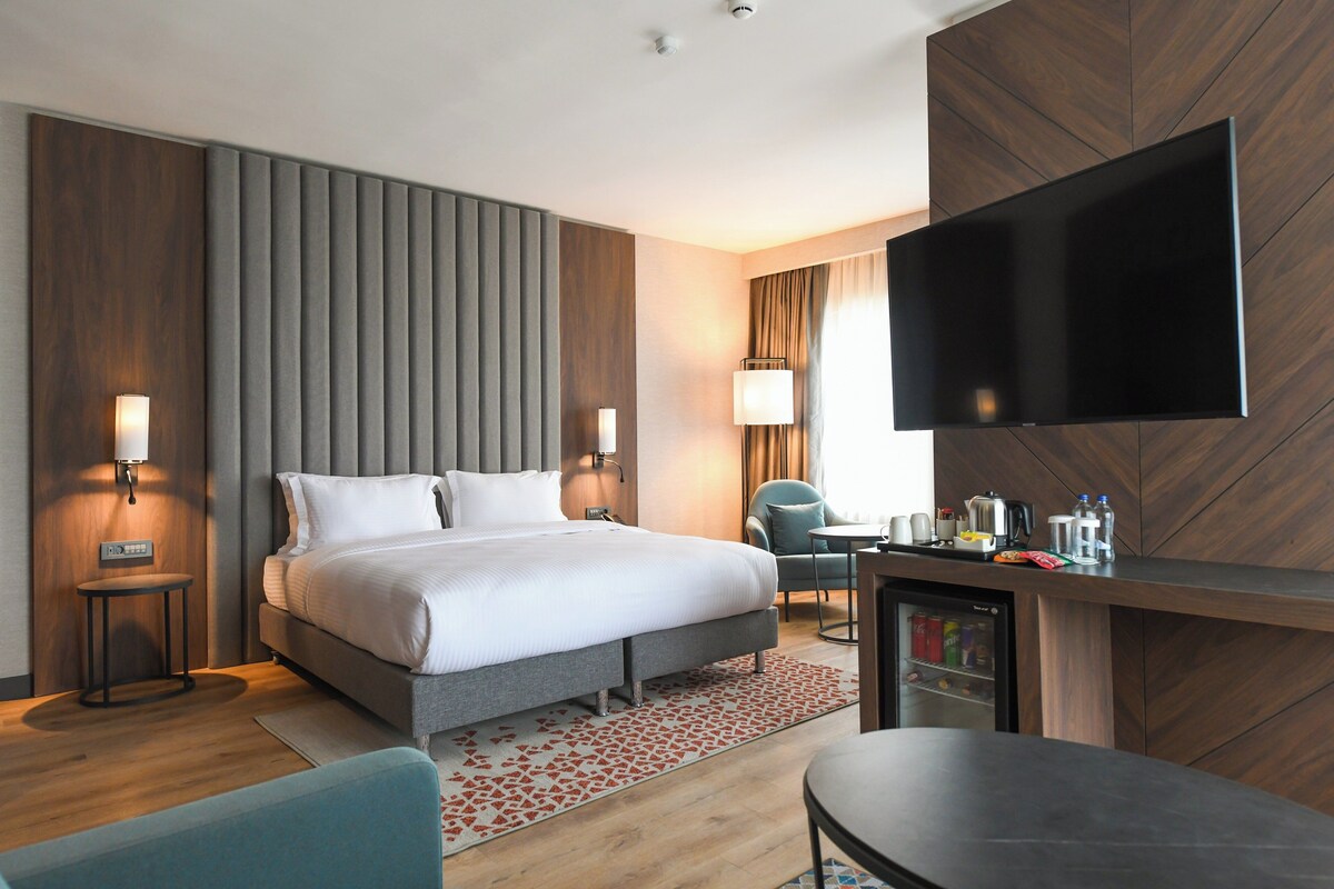 A modern hotel room with a bed, bedside tables, a TV, a desk, and a mini-fridge.