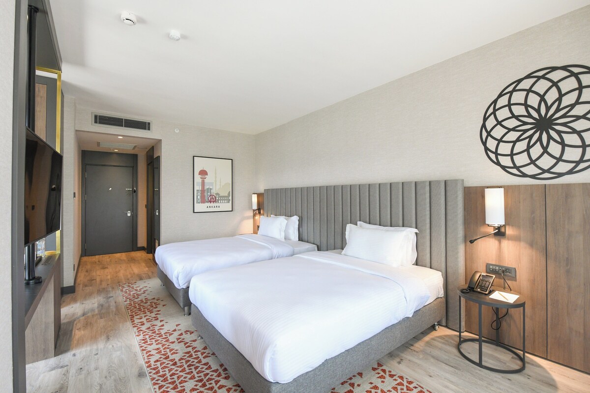 A hotel room with two beds, a TV, a bedside table with a phone, and a decorative wall piece.