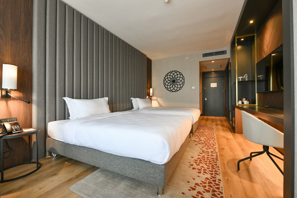 A modern hotel room with two beds, a desk, a chair, and a bathroom visible through an open door.