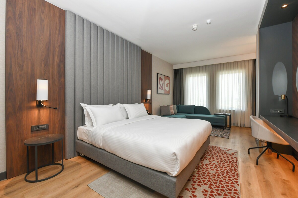 A modern hotel room with a large bed, a desk, a sofa, and a view of the city.