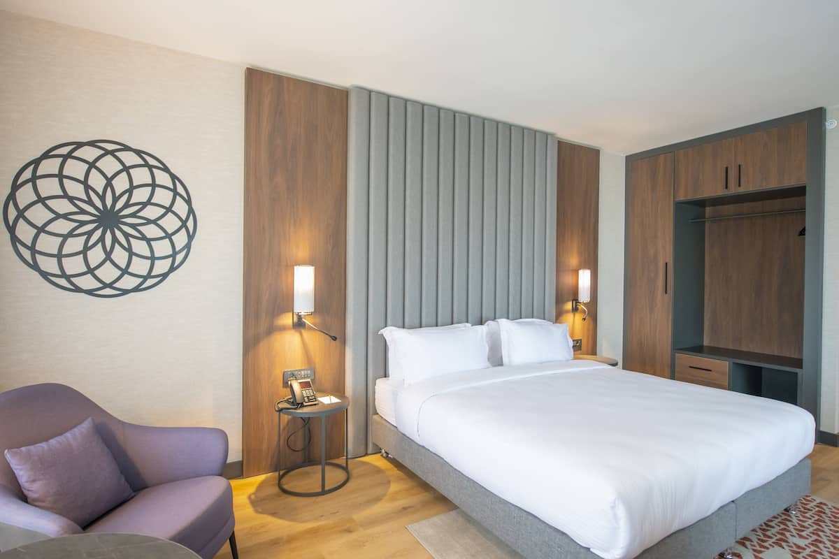 A modern hotel room with a large bed, a chair, a desk with a phone, and a wardrobe.