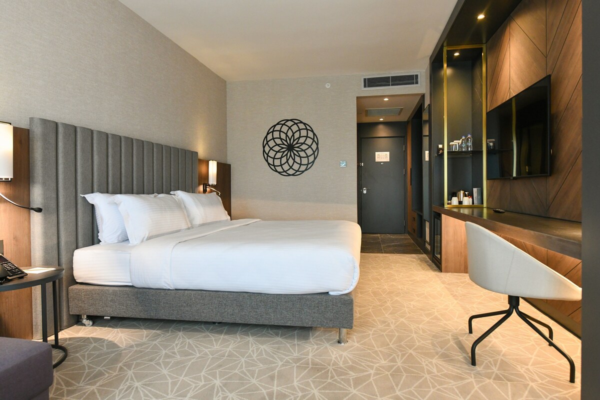 A modern hotel room with a large bed, a desk with a lamp, a chair, a TV, and a decorative wall piece.