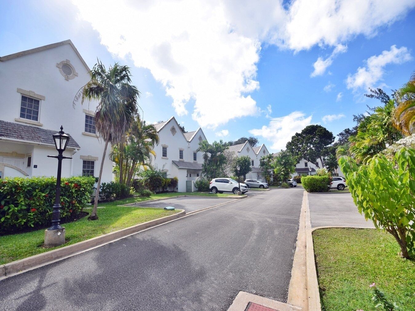 Una calle residencial con casas blancas, coches aparcados y abundante vegetación.