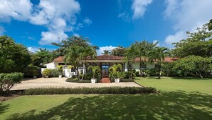 Una casa de dos pisos con techo de tejas, rodeada de un césped cuidado y diversas plantas tropicales.