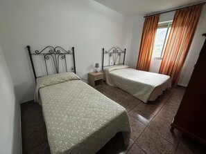 Two single beds with metal headboards and matching bedspreads, a bedside table with a lamp, and a window with striped curtains.