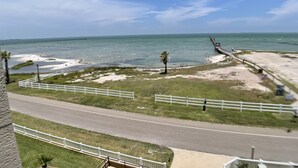 Eine Küstenstraße mit weißem Zaun, einer Palme und Blick auf das Meer und einen Steg.