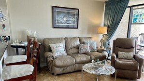 Ein Wohnzimmer mit einem beigen Sofa, zwei Sesseln, einem Couchtisch und einem Essbereich mit Stühlen und einem Tisch.