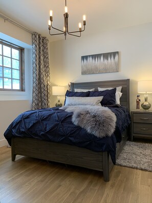Ein Schlafzimmer mit einem dunkelholznen Bett, gemusterten Vorhängen, einer Chandelieber, einem Gemälde an der Wand und einem Nachttisch mit Lampe.
