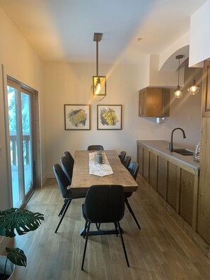 Eine moderne Küche mit einem Holztisch, schwarzen Stühlen und einer zentralen Leuchte. Zwei gerahmte Kunstwerke hängen an der Wand, und ein Fenster bietet Blick auf Bäume.