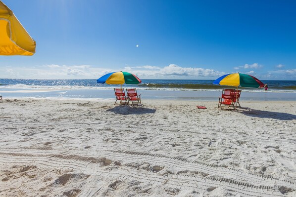A beach with two red beach chairs under colorful umbrellas, a clear sky, and the ocean in the background. - "Sunshine At The Beach - Fall Specials! Many Upgrades! Perfect Location! (Orange Beach)