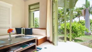 Una habitación con una mesa de vidrio, un sofá con cojines, una ventana con contraventanas y vistas a palmeras y follaje exterior.
