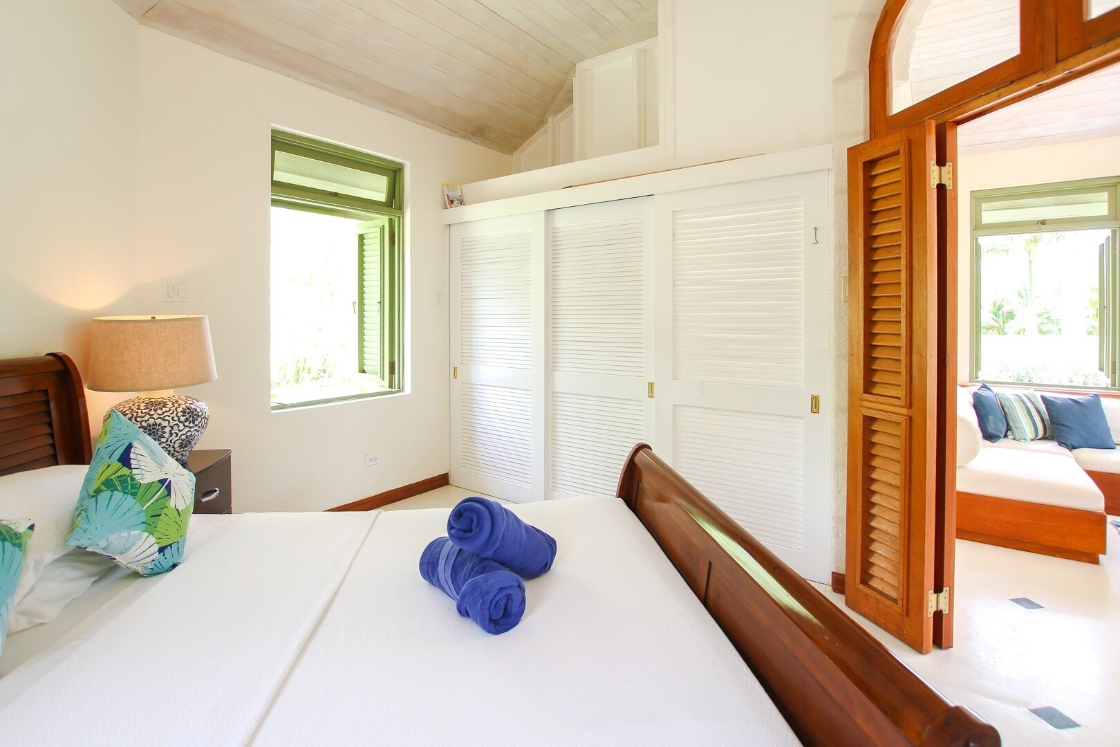 A bedroom with a bed, a nightstand, a lamp, a window, and a set of white louvered doors.