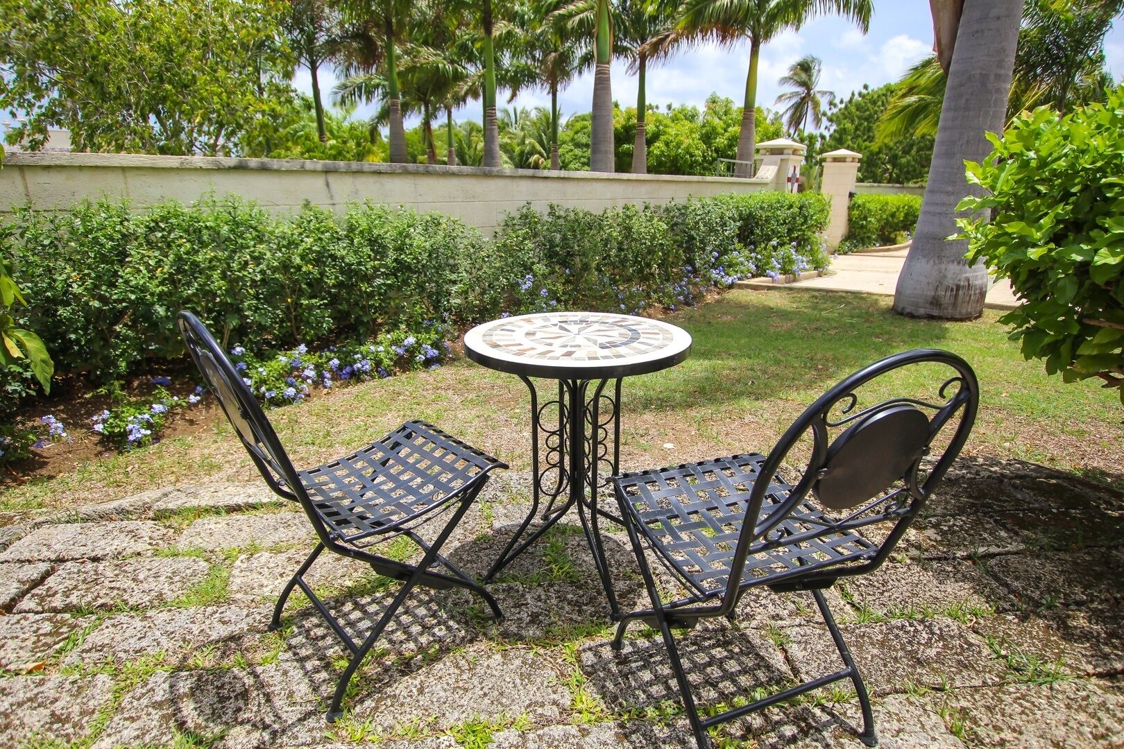 A patio with a round table and two chairs, surrounded by greenery and palm trees.