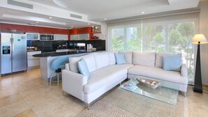 A modern kitchen with a central island, a white sofa, and a large window with a view of greenery. - Stunning Beachfront 2-Bed Condo with Pool - Ocean One 204 by BSL Rentals (Maxwell)
