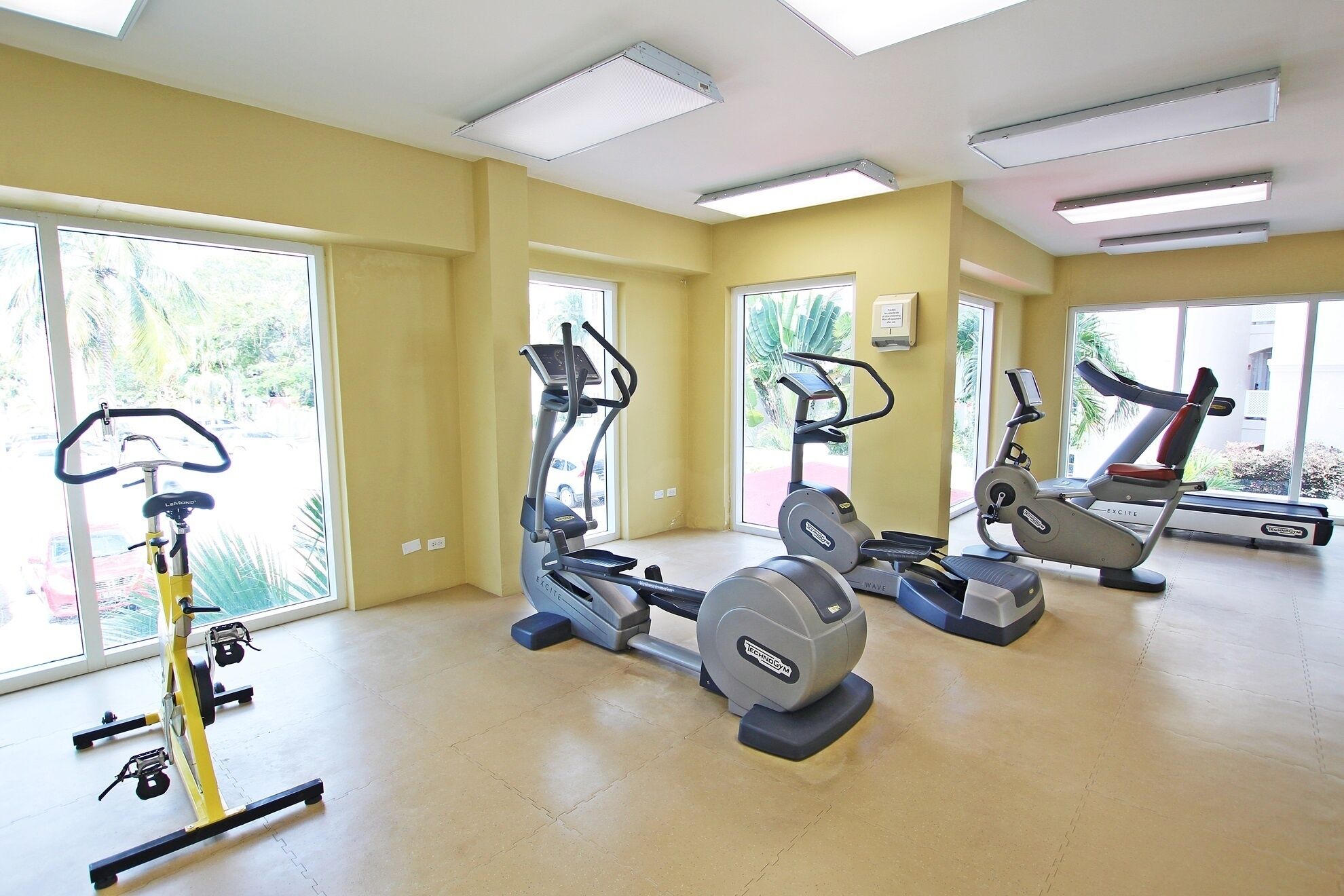 A gym with multiple exercise machines, large windows, and a yellow wall.