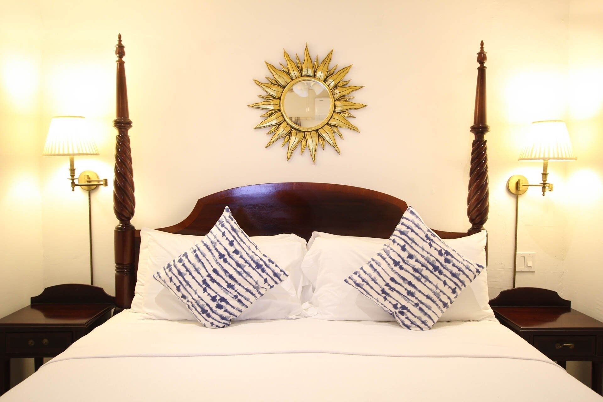 A neatly made bed with white and blue patterned pillows, a wooden headboard, and a sunburst mirror on the wall.
