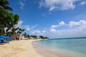 A beach with clear turquoise water, palm trees, and a row of beach chairs and umbrellas. - Poolside Paradise Near Beach - Pavilion Villa by Bluescape (Holetown)