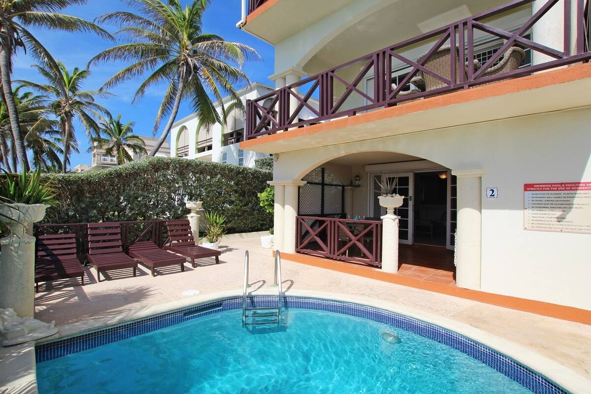 A pool area with a clear blue pool, wooden lounge chairs, and a building with a balcony.