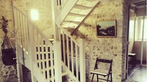 A staircase with white railings leading to a landing. A chair is placed on the landing, and a painting hangs on the wall. - Normandieheritage14 Cottage du Marais Omaha Beach D-Day Tour Guide E-bike Hiking (Canchy)