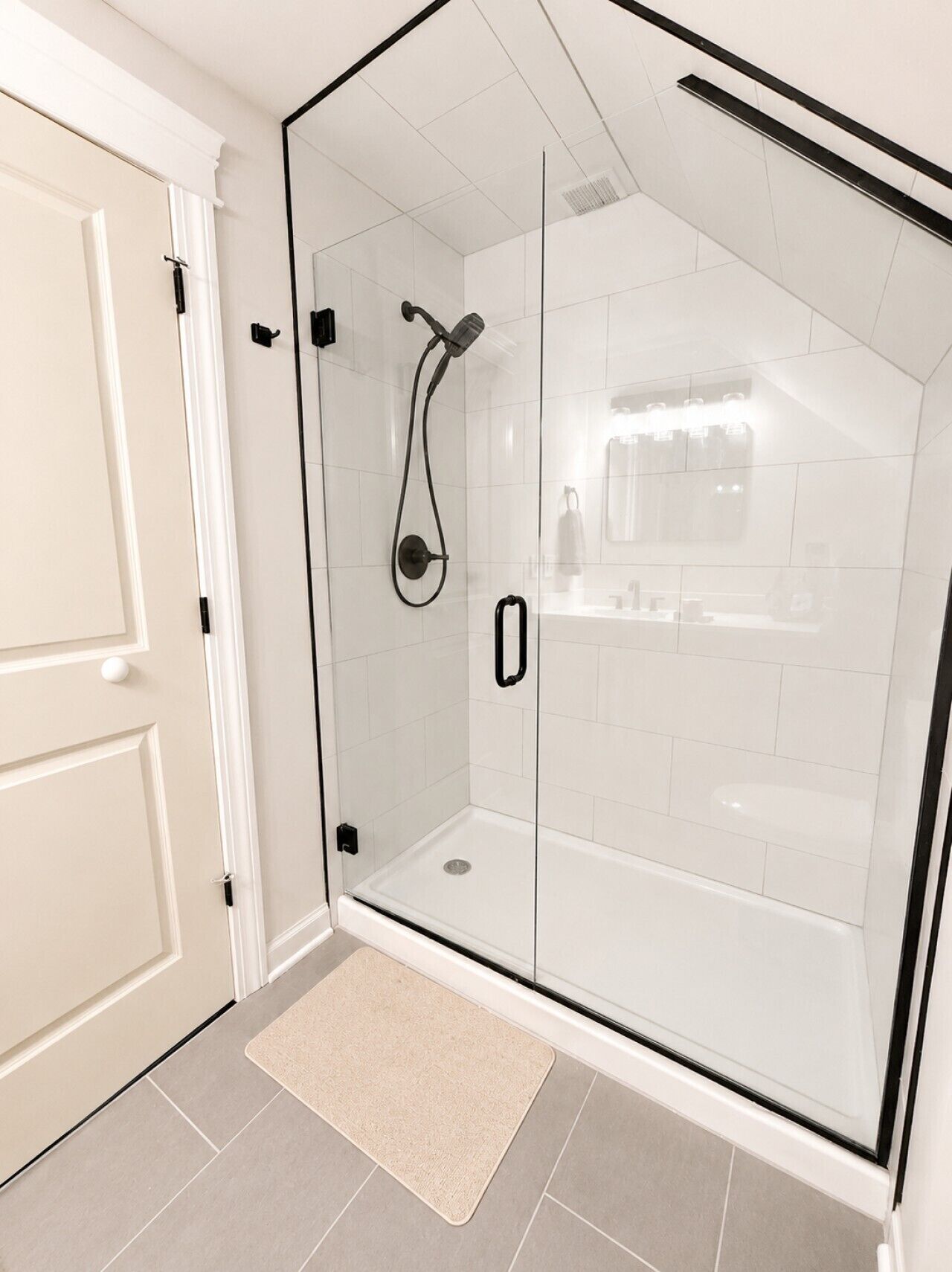 A glass-enclosed shower with a handheld showerhead, a white door, and a beige rug on a tiled floor.