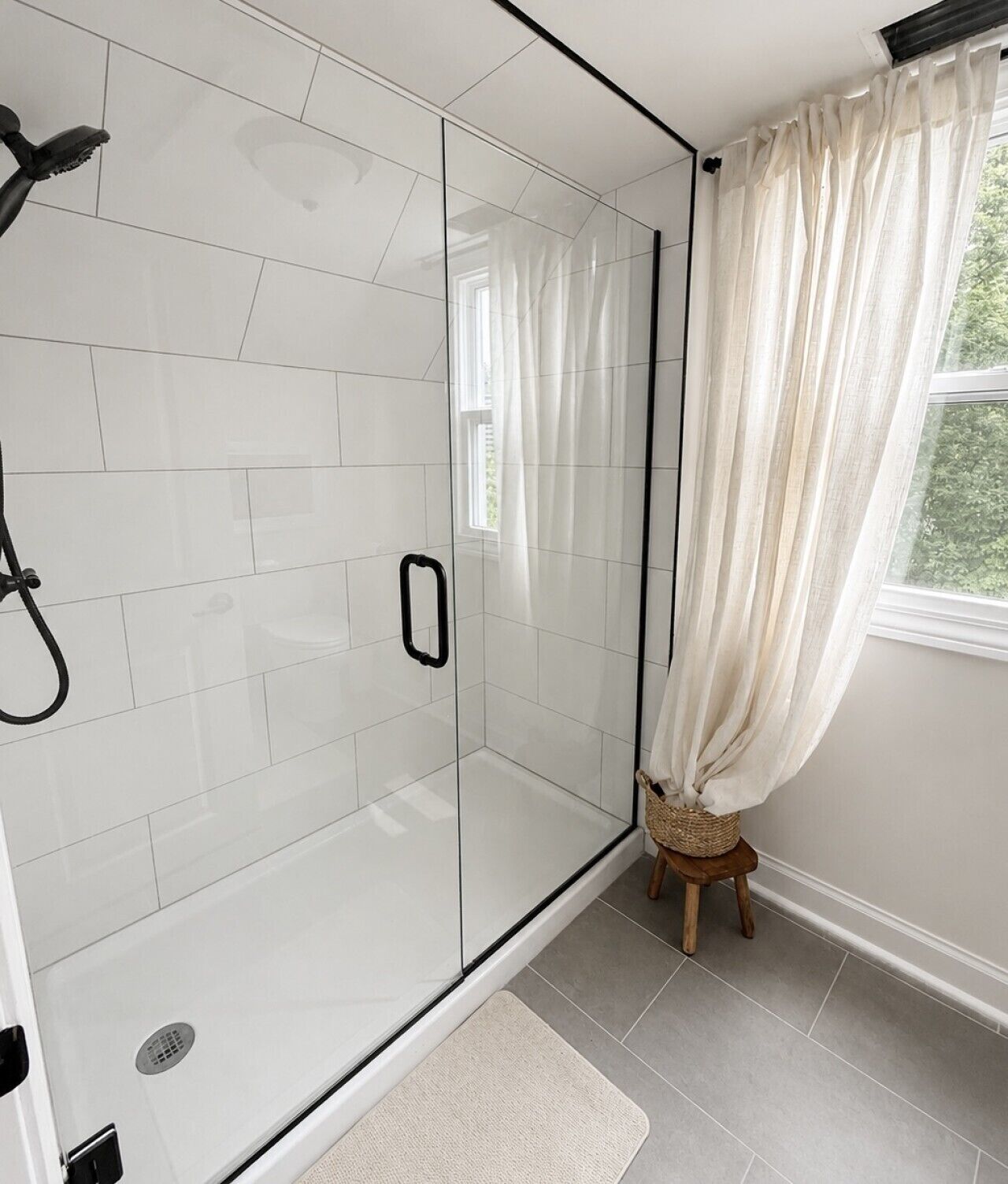 A modern bathroom with a glass shower enclosure, white tiled walls, a wooden stool, and a window with sheer curtains.