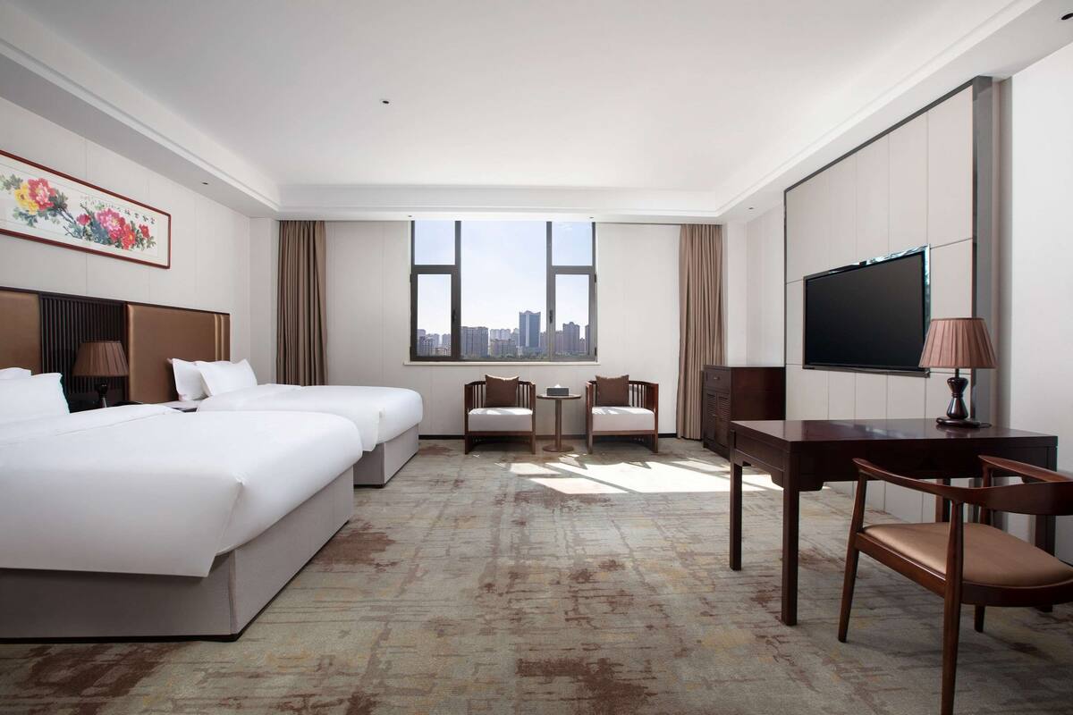 A hotel room with two beds, a desk, a chair, a TV, and a window with a city view.