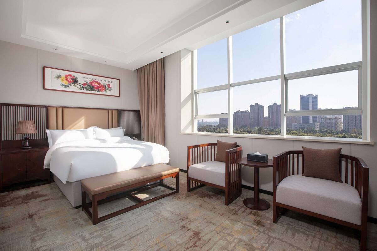 A hotel room with a bed, a desk, two chairs, a window with a city view, and a framed artwork on the wall.