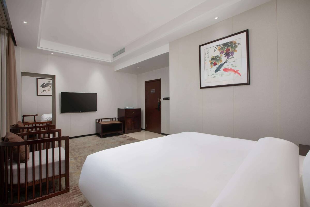 A hotel room with a large bed, a TV mounted on the wall, a wooden crib, a desk, and a wardrobe.
