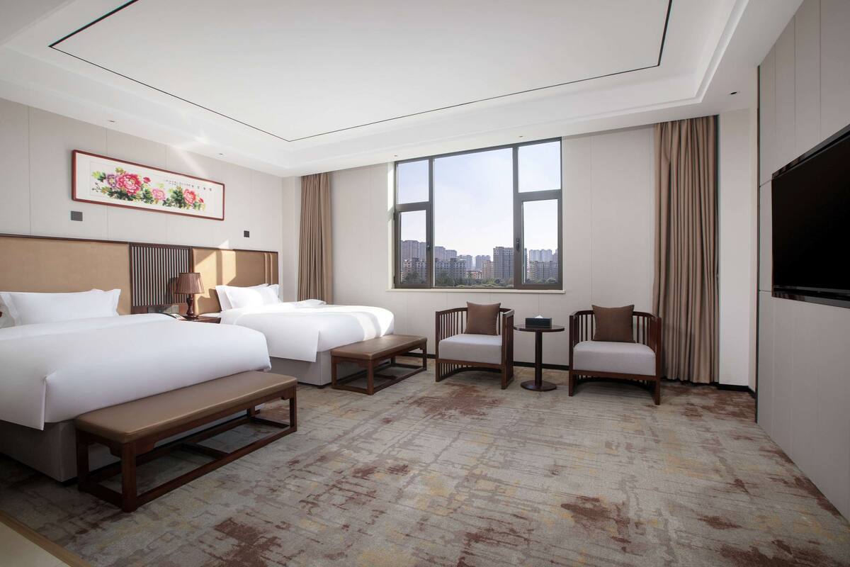 A hotel room with two beds, a desk, chairs, a large window, and a city view.
