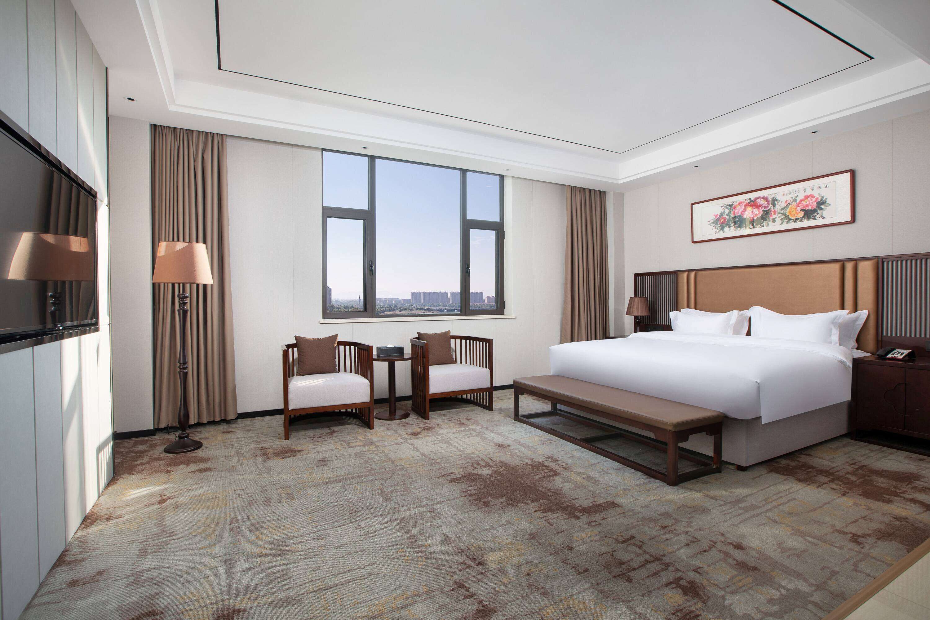 Habitación de hotel con una cama grande, un escritorio con dos sillas, vistas de la ciudad a través de amplios ventanales y un cuadro en la pared.