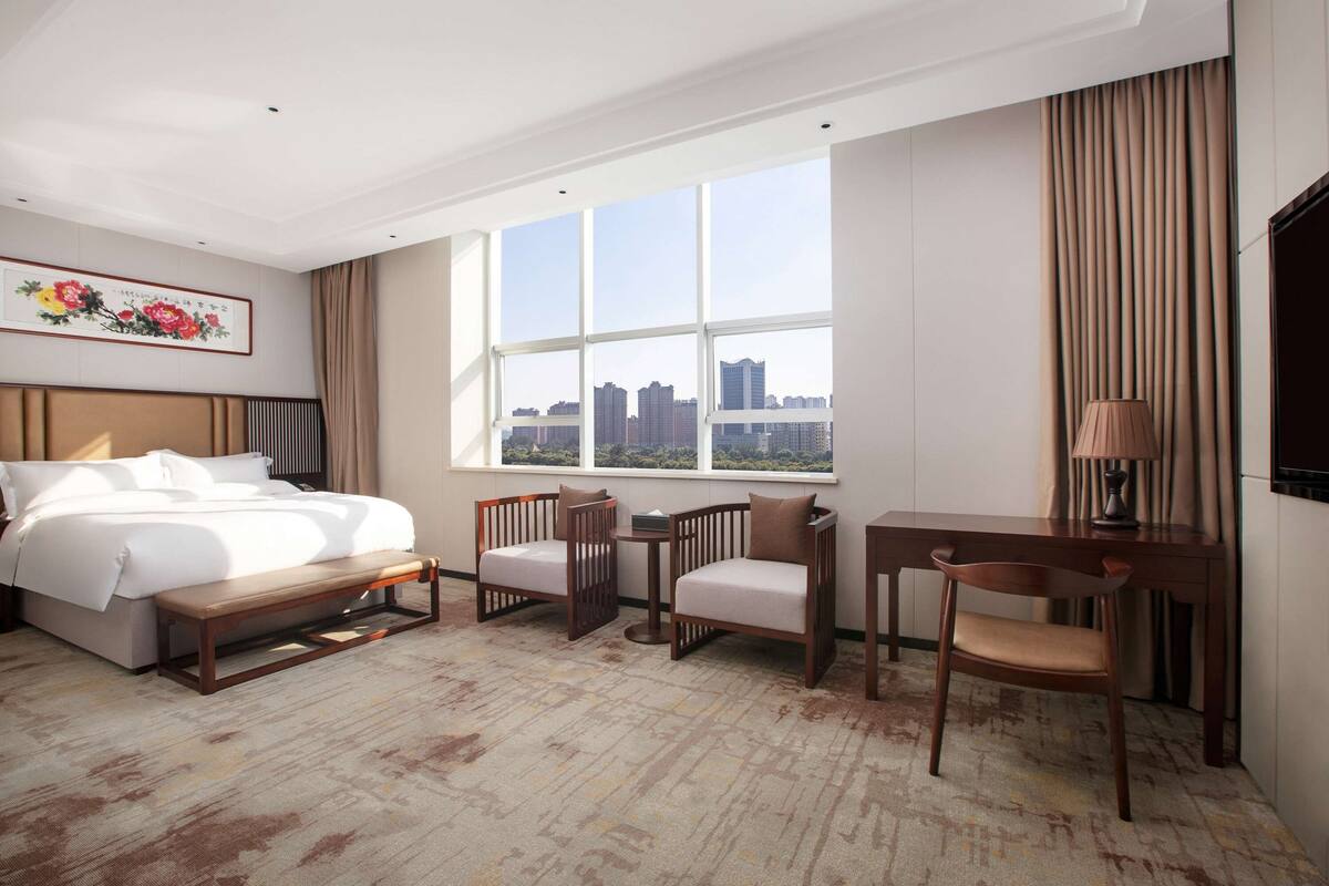 A hotel room with a bed, two chairs, a desk, a bench, and a large window with a city view.