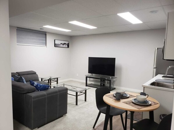 Um espaço de estar compacto com sofá, televisão sobre suporte, mesa pequena com cadeiras e área de cozinha compacta.