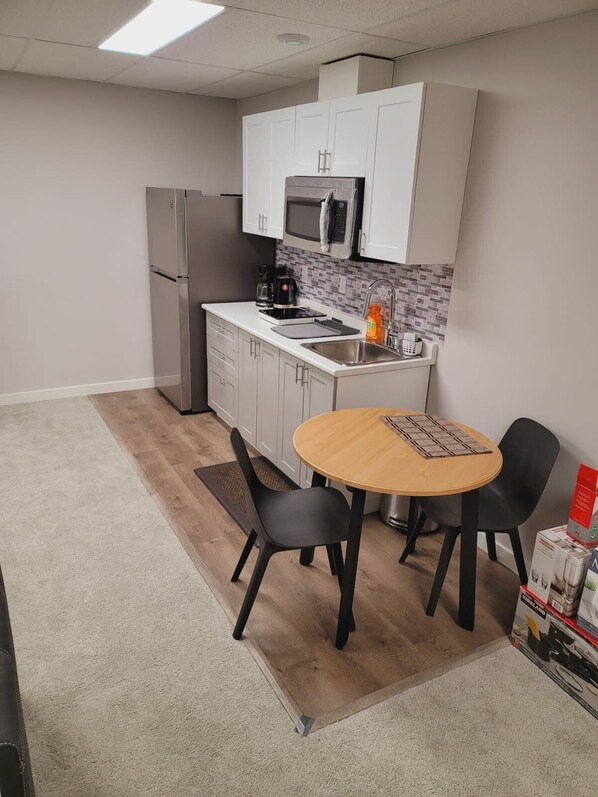 A small kitchenette with a refrigerator, microwave, and coffee maker. There is a round wooden table with two chairs and a small rug. - Cozy 2 bedroom basement (Winnipeg)