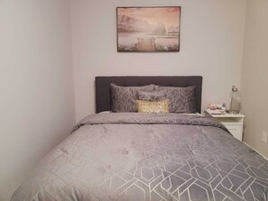 Quarto com cama, cabeceira, mesa de cabeceira com abajur e um quadro na parede.