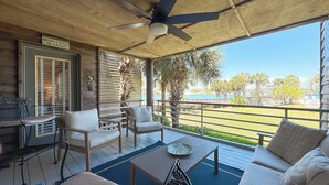 Ein überdachter Außenbereich mit Sitzgelegenheit, Deckenventilator, Tisch mit dekorativer Platte und Blick auf Strand und Palmen.