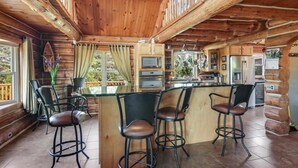 A rustic cabin interior with a wooden ceiling, a central island with bar stools, a kitchen area, and large windows.