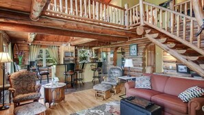 A spacious log cabin interior with a wooden staircase, a cozy living area with a sofa and armchairs, and a kitchen in the background.