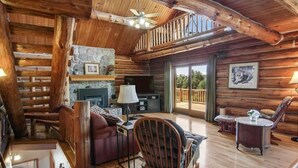 A log cabin interior with a fireplace, wooden furniture, and a balcony with a view.