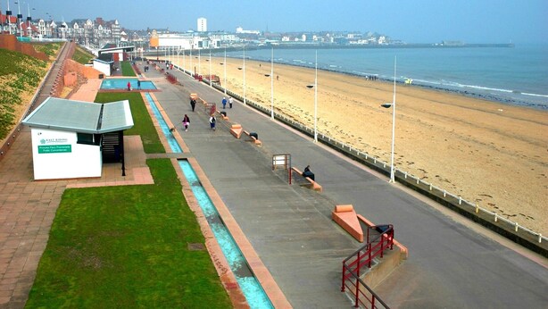 Une promenade en bord de mer avec une plage, une allée piétonne et un bâtiment portant l’inscription « Toilettes publiques de la promenade East Riding Princess Mary ».