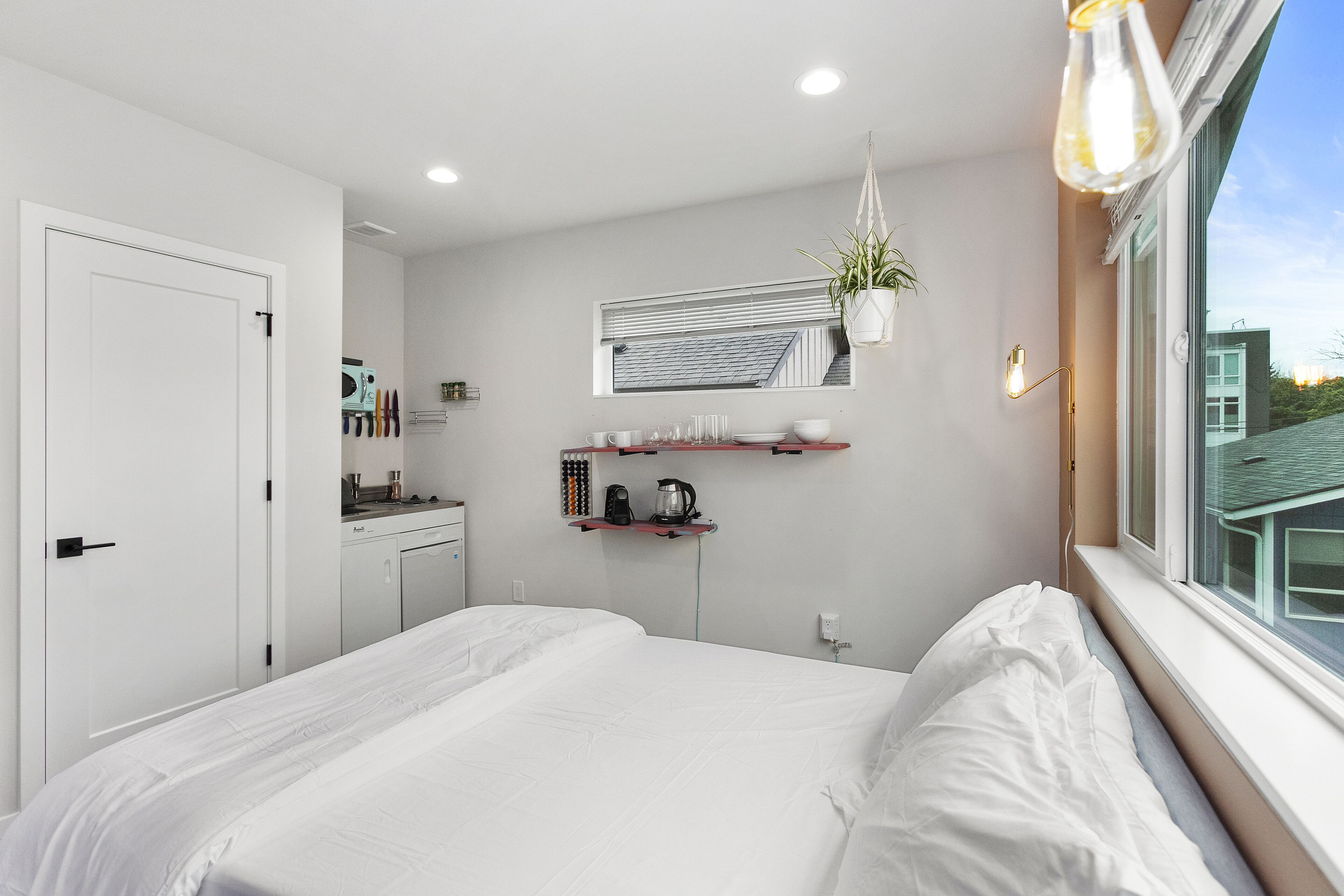 A modern bedroom with a large bed, a small kitchenette, a window with a view, and a hanging plant.