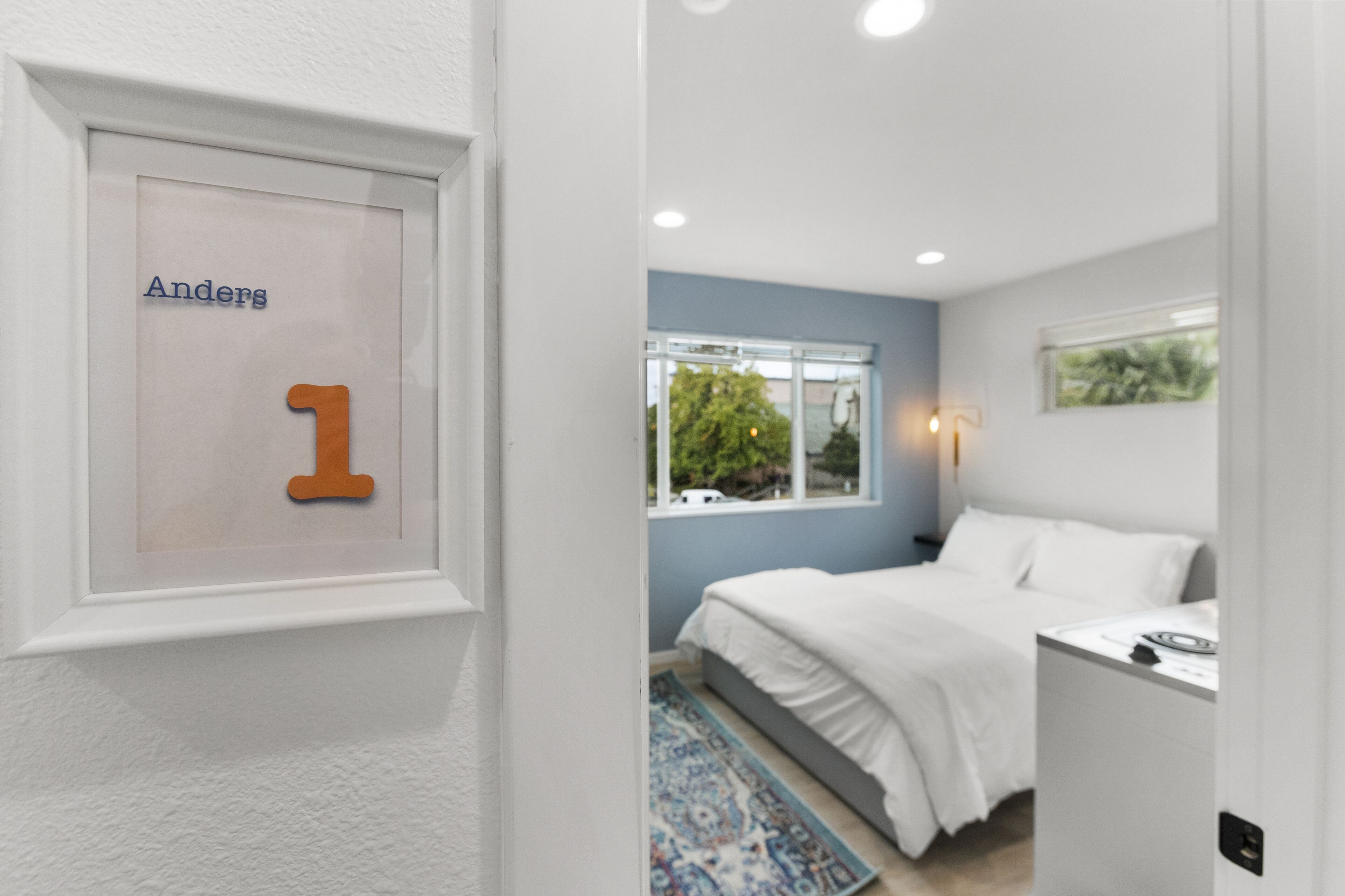 A bedroom with a bed, a window, a bedside table, and a wall-mounted number sign.