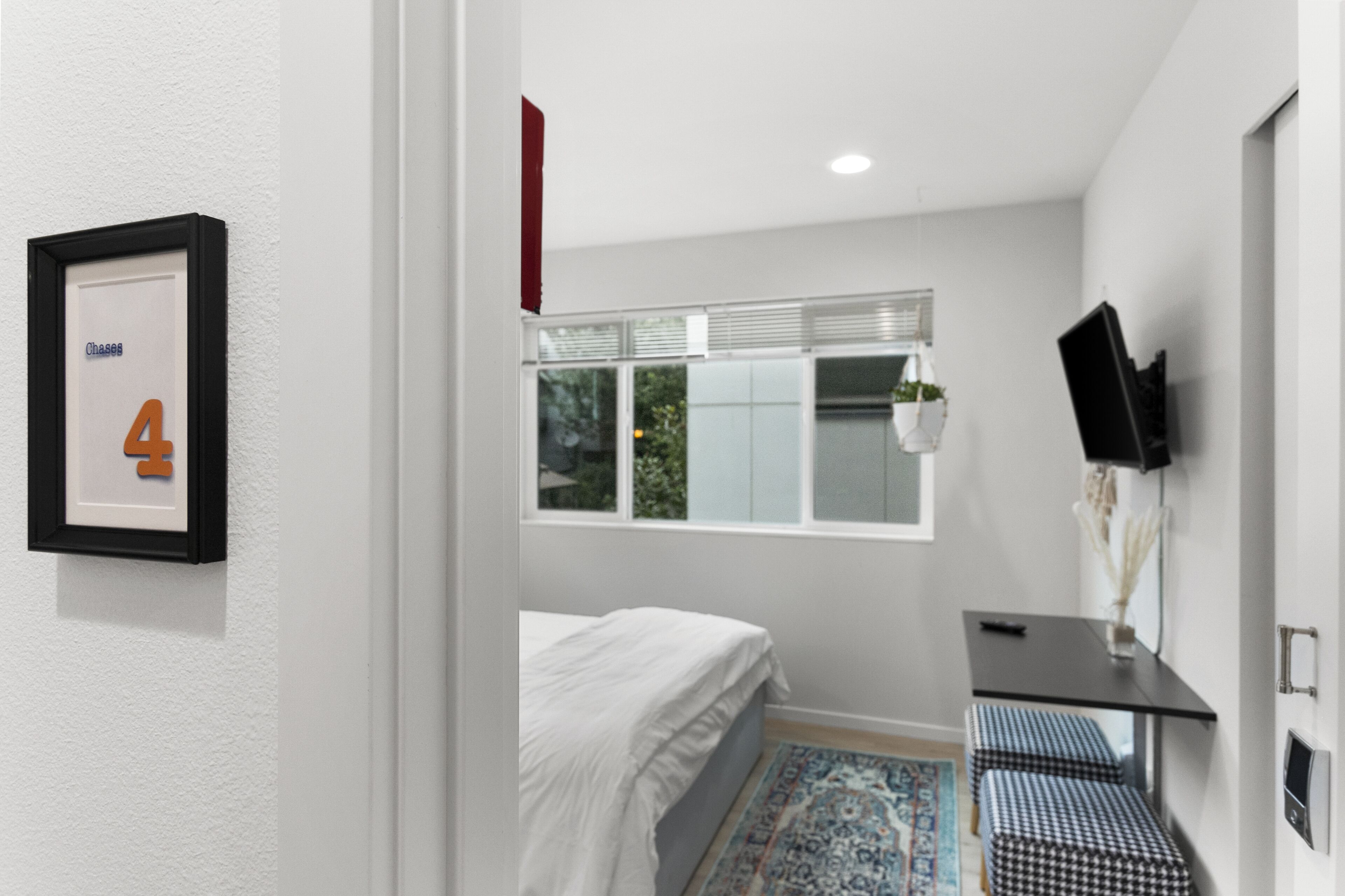 A bedroom with a bed, a desk, a chair, a TV, and a window with blinds.